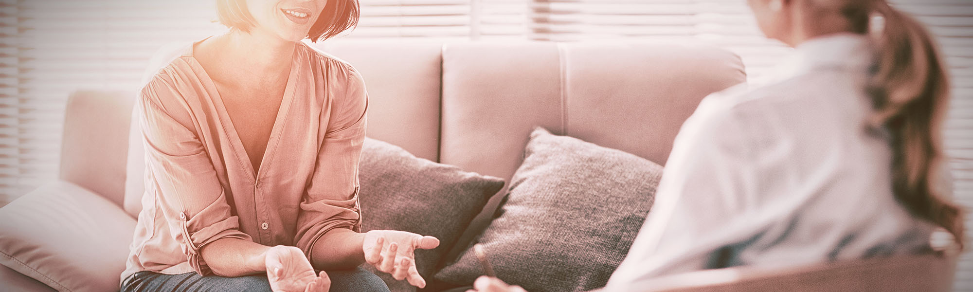 Zwei Frauen sitzen sich gegenüber in einem hellem Wohnzimmer: Die Beraterin im Vordergrund (nur von hinten zu sehen) trägt eine weiße Bluse und spricht mit der Klientin auf dem Sofa. Die Klientin, eine lächelnde Frau mit kastanienfarbenem Bob und rosafarbenem Oberteil, gestikuliert offen mit Händen, während sie auf hellem Polstersofa mit Kissen sitzt. Der Raum ist sanft von Tageslicht durch Fensterjalousien erhellt und vermittelt eine freundliche Beratungssituation.